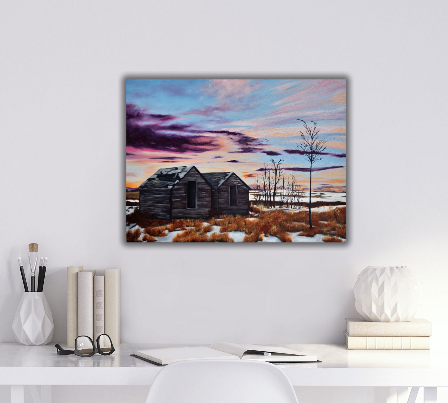 White desk with stationary and glasses on it, and a landscape painting of a purple and orange sunset with old sheds on the wall