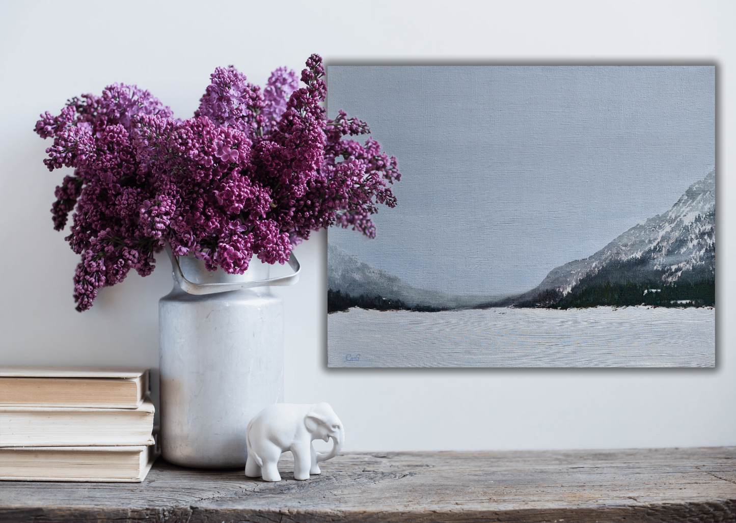 A wood table with books and purple lilacs in a pot with a landscape painting of mountains and a frozen lake in fog in the background